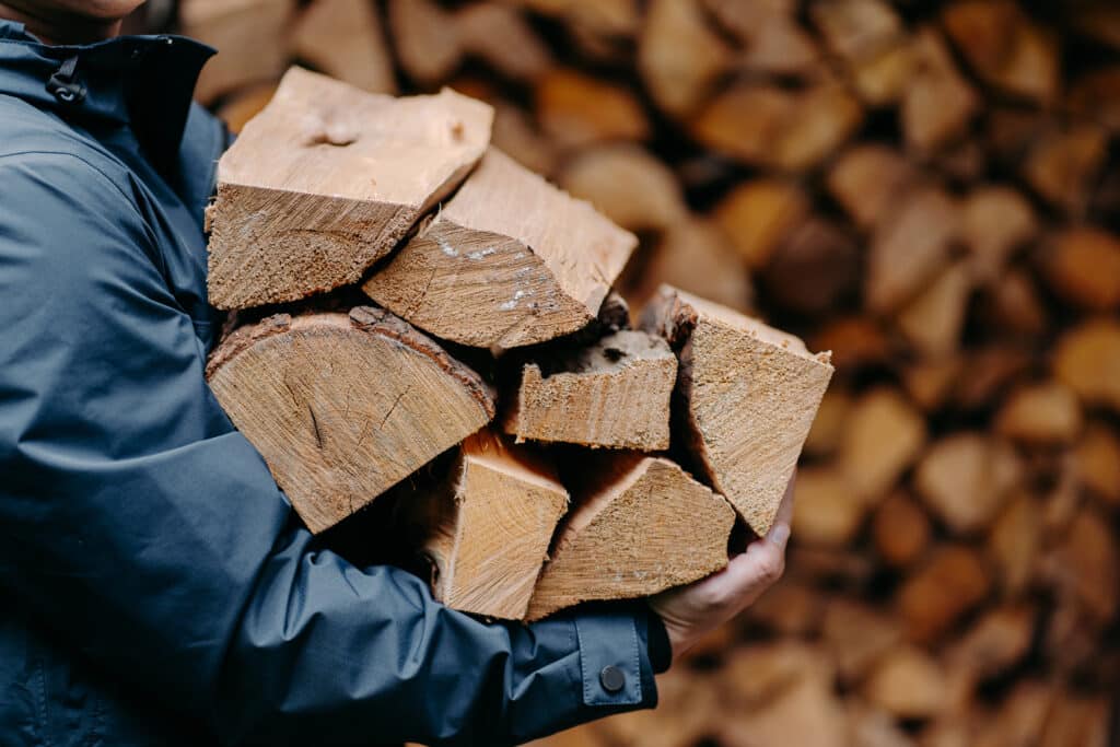 Homme en veste tient un grand tas de bûches de bois de chauffage devant un stock empilé, prêt pour l'hiver.