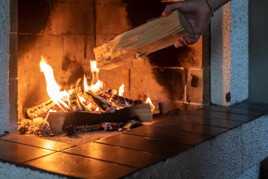 Main posant une bûche de bois dans une cheminée en briques. Un feu ardent avec des flammes oranges illumine l'âtre.